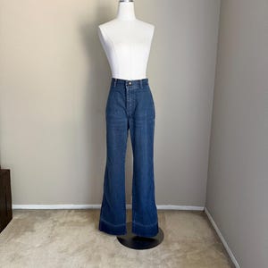 K&ouml;nnte beinhalten: Dunkelblaue Jeanshose mit Schlaghose, pr&auml;sentiert auf einer wei&szlig;en Schaufensterpuppe. Die Hose hat eine hohe Taille, einen Knopf- und Rei&szlig;verschluss und ein ausgestelltes Bein-Design. Die Schaufensterpuppe steht auf einem schwarzen St&auml;nder vor einer neutralfarbenen Wand und Teppich.