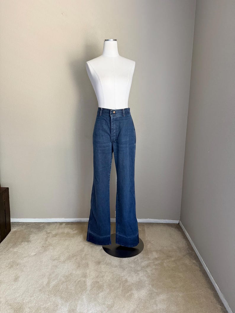 K&ouml;nnte beinhalten: Dunkelblaue Jeanshose mit Schlaghose, pr&auml;sentiert auf einer wei&szlig;en Schaufensterpuppe. Die Hose hat eine hohe Taille, einen Knopf- und Rei&szlig;verschluss und ein ausgestelltes Bein-Design. Die Schaufensterpuppe steht auf einem schwarzen St&auml;nder vor einer neutralfarbenen Wand und Teppich.