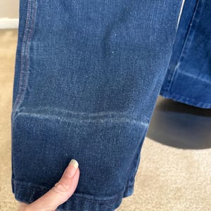 K&ouml;nnte beinhalten: Nahaufnahme einer dunkelblauen Jeans. Die Jeans hat ein klassisches Design mit sichtbarer Naht und Saum am unteren Ende. Der Stoff weist subtile Abnutzungserscheinungen auf, was dem Kleidungsst&uuml;ck Charakter verleiht. Die Jeans wird von einer Hand gehalten.
