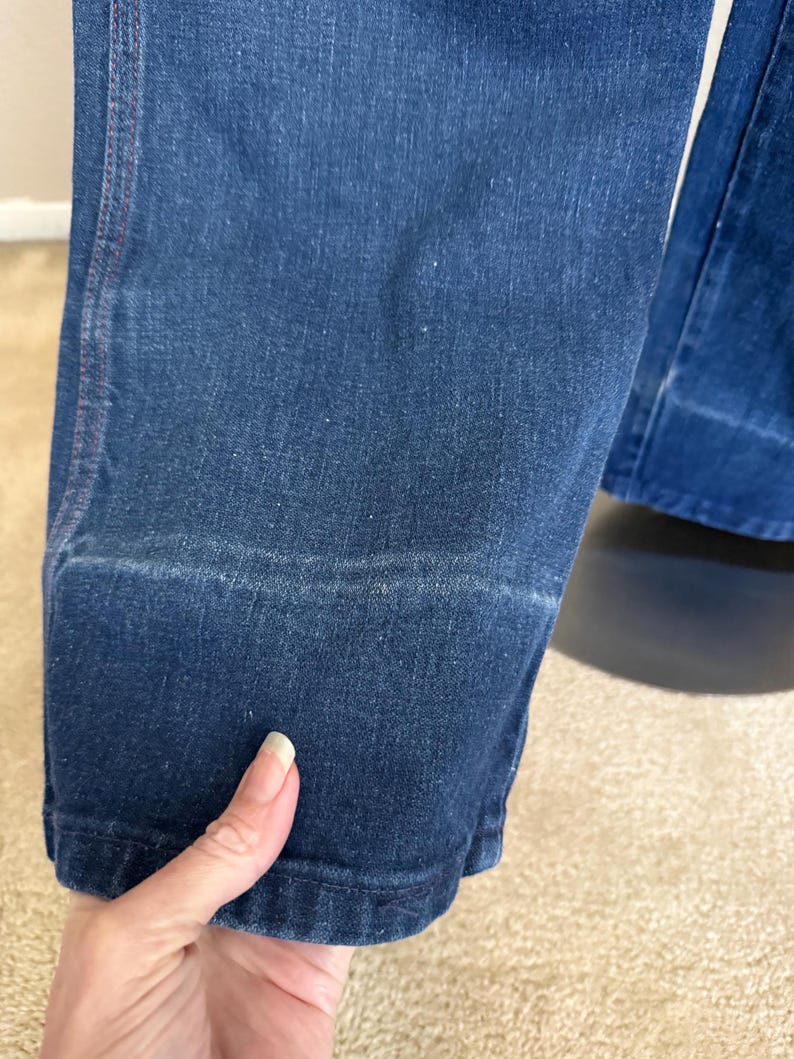 K&ouml;nnte beinhalten: Nahaufnahme einer dunkelblauen Jeans. Die Jeans hat ein klassisches Design mit sichtbarer Naht und Saum am unteren Ende. Der Stoff weist subtile Abnutzungserscheinungen auf, was dem Kleidungsst&uuml;ck Charakter verleiht. Die Jeans wird von einer Hand gehalten.
