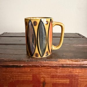 May include: A ceramic mug with a yellow base color featuring a repeating pattern of vertical, elongated diamond shapes in shades of dark brown, black, and orange, accented by small brown circular dots between the shapes and a curved handle.
