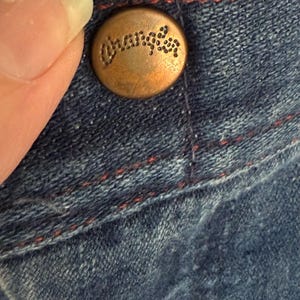 K&ouml;nnte beinhalten: Nahaufnahme eines kupferfarbenen Knopfes mit dem eingravierten Wort "Wrangler". Der Knopf ist an blauem Denim-Stoff mit roten N&auml;hten befestigt. Der Denim hat ein abgenutztes Aussehen.