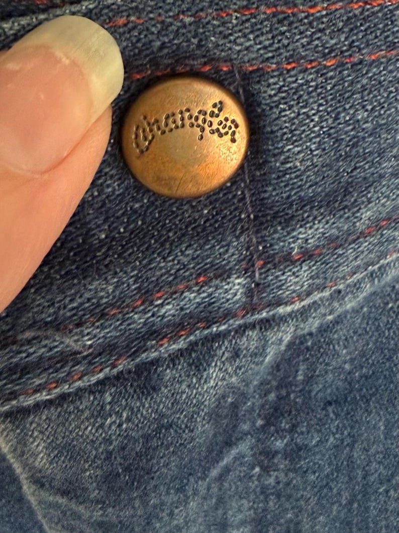 K&ouml;nnte beinhalten: Nahaufnahme eines kupferfarbenen Knopfes mit dem eingravierten Wort "Wrangler". Der Knopf ist an blauem Denim-Stoff mit roten N&auml;hten befestigt. Der Denim hat ein abgenutztes Aussehen.