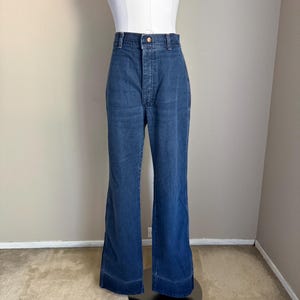 K&ouml;nnte beinhalten: Blaue Jeanshose mit Schlaghose. Die hoch taillierte Hose hat einen Knopf- und Rei&szlig;verschluss. Die Hose wird auf einem wei&szlig;en Mannequin-Oberk&ouml;rper pr&auml;sentiert und steht auf einem schwarzen Sockel. Die Hose hat eine ausgestellte Bein&ouml;ffnung.