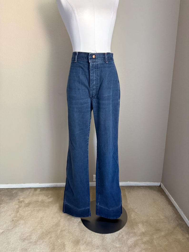 K&ouml;nnte beinhalten: Blaue Jeanshose mit Schlaghose. Die hoch taillierte Hose hat einen Knopf- und Rei&szlig;verschluss. Die Hose wird auf einem wei&szlig;en Mannequin-Oberk&ouml;rper pr&auml;sentiert und steht auf einem schwarzen Sockel. Die Hose hat eine ausgestellte Bein&ouml;ffnung.