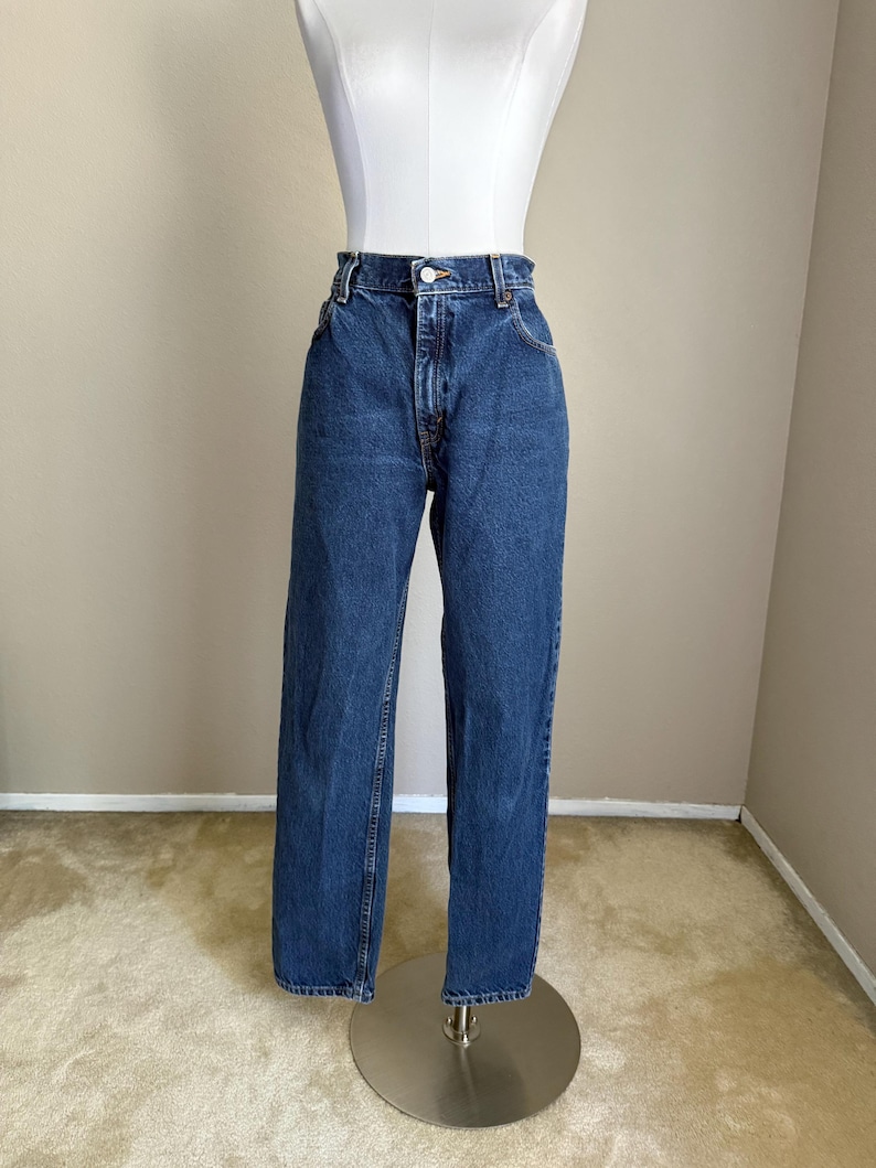 May include: High-waisted blue denim jeans featuring a classic five-pocket design, a metal button closure, and a zipper fly. The jeans have a straight-leg cut with a medium wash finish, visible orange stitching, and belt loops along the waistband.