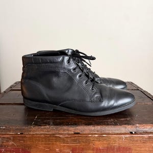 May include: A pair of black leather ankle boots with laces. The boots are sitting on a wooden crate. The boots have a rounded toe and a low heel. The laces are black and tied in a bow.