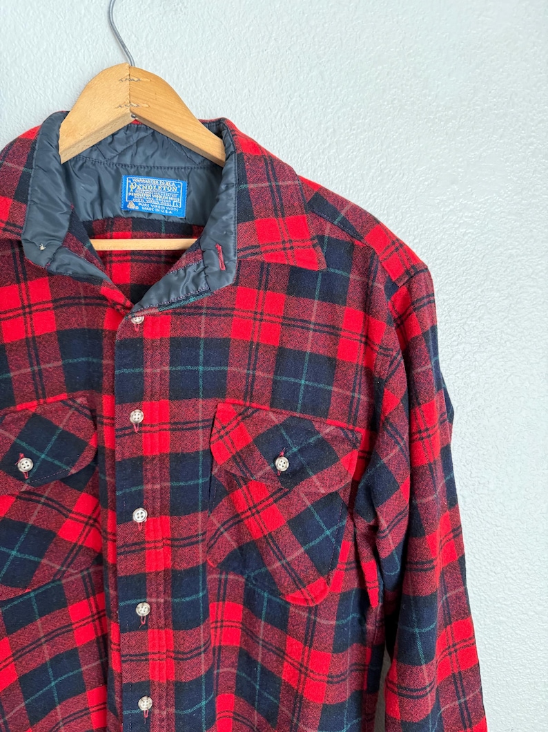 Puede incluir: Camisa de franela a cuadros rojos y negros con dos bolsillos en el pecho y cuello azul marino acolchado. La camisa es abotonada con botones plateados y una etiqueta que dice "Pendleton". Colgada en una percha de madera.