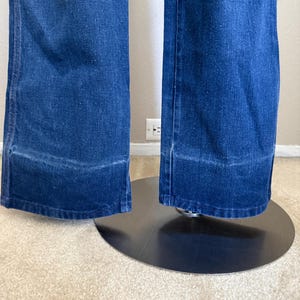 K&ouml;nnte beinhalten: Nahaufnahme einer blauen Jeanshose mit weitem Bein. Die Hose hat einen klassischen F&uuml;nf-Taschen-Stil und eine mittelblaue Waschung. Der Saum ist mit einer sauberen Kante versehen. Die Hose wird auf einem schwarzen St&auml;nder pr&auml;sentiert.