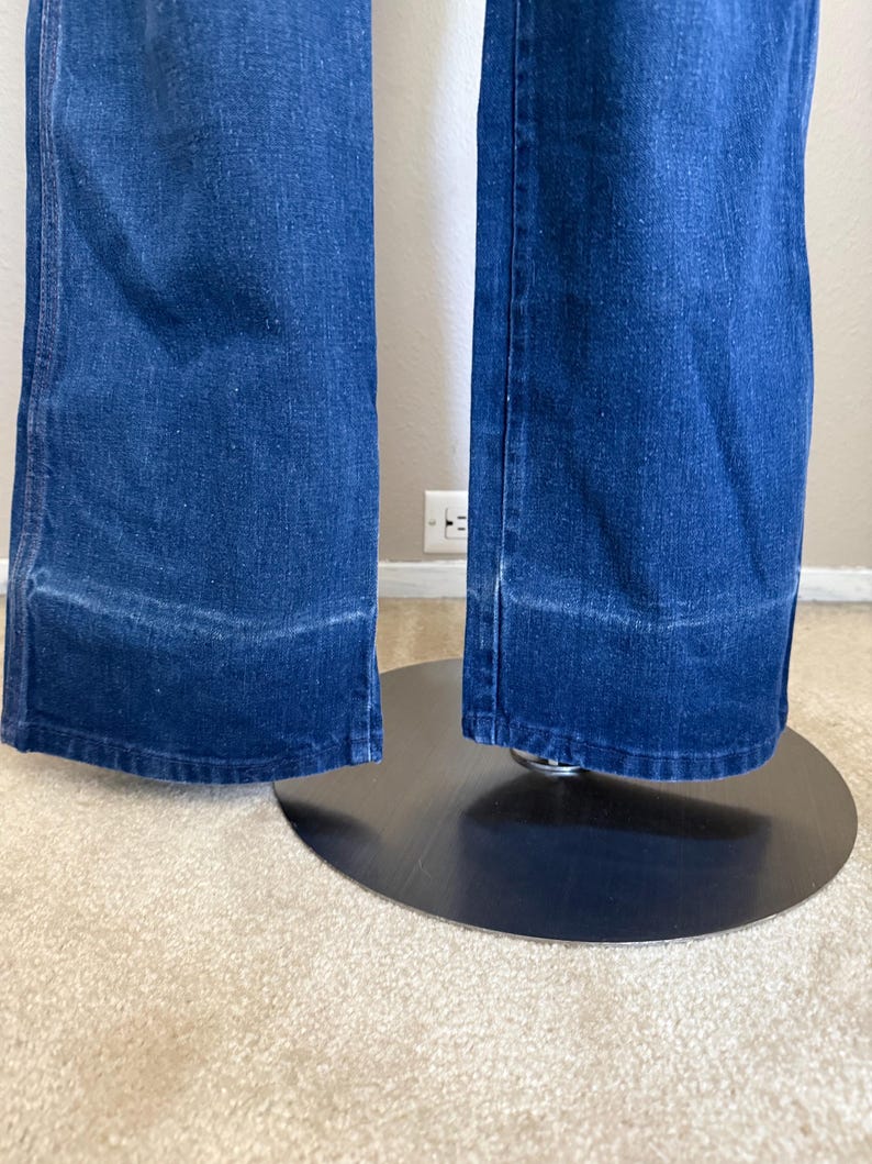 K&ouml;nnte beinhalten: Nahaufnahme einer blauen Jeanshose mit weitem Bein. Die Hose hat einen klassischen F&uuml;nf-Taschen-Stil und eine mittelblaue Waschung. Der Saum ist mit einer sauberen Kante versehen. Die Hose wird auf einem schwarzen St&auml;nder pr&auml;sentiert.