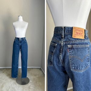 May include: Dark blue denim jeans featuring a high-waisted fit, a button and zipper fly closure, and belt loops. The back includes a tan leather-like patch with the text Levi Strauss & Co. Quality Clothing 550 and a small red brand tab.