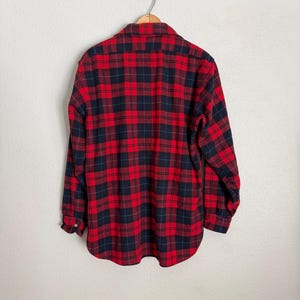 Puede incluir: Camisa de franela a cuadros rojos y negros colgada de una percha de madera. La camisa de manga larga tiene un dise&ntilde;o cl&aacute;sico con botones, cuello y pu&ntilde;os abotonados. La tela presenta un patr&oacute;n a cuadros llamativo.