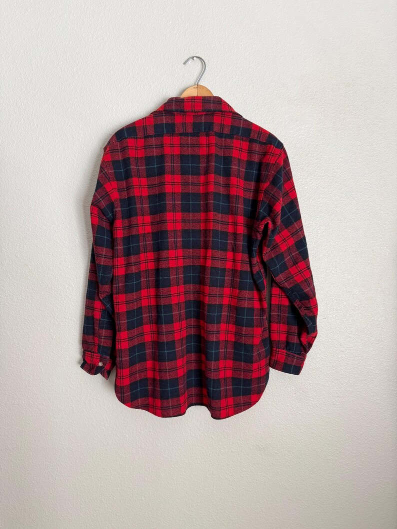 Puede incluir: Camisa de franela a cuadros rojos y negros colgada de una percha de madera. La camisa de manga larga tiene un dise&ntilde;o cl&aacute;sico con botones, cuello y pu&ntilde;os abotonados. La tela presenta un patr&oacute;n a cuadros llamativo.