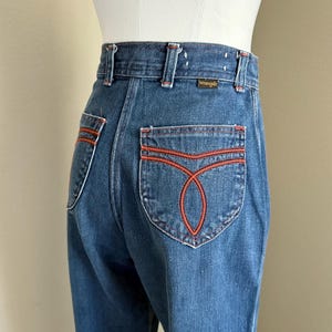 K&ouml;nnte beinhalten: Dunkelblaue Wrangler Jeans mit orangefarbenen N&auml;hten. Diese Jeans mit hohem Bund hat ein gerades Bein und dekorative N&auml;hte auf den Ges&auml;&szlig;taschen. Das Wrangler-Logo ist auf dem Bund zu sehen.