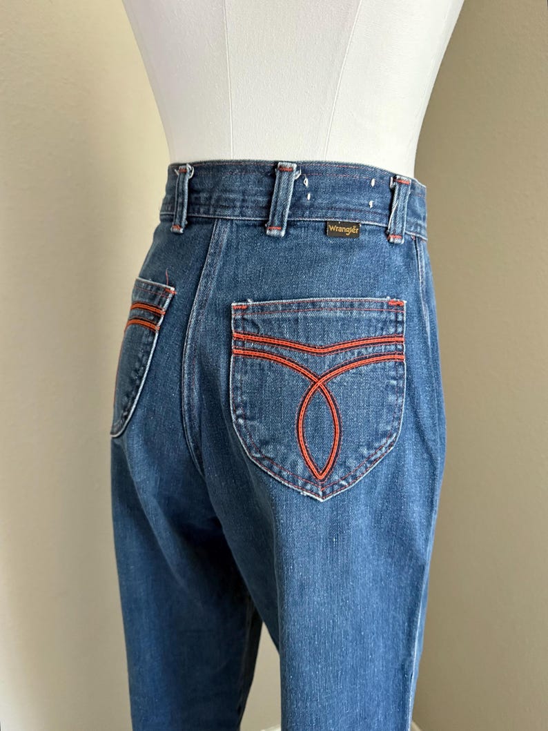 K&ouml;nnte beinhalten: Dunkelblaue Wrangler Jeans mit orangefarbenen N&auml;hten. Diese Jeans mit hohem Bund hat ein gerades Bein und dekorative N&auml;hte auf den Ges&auml;&szlig;taschen. Das Wrangler-Logo ist auf dem Bund zu sehen.