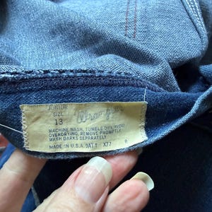 K&ouml;nnte beinhalten: Nahaufnahme einer blauen Jeans mit einem Wrangler-Etikett. Das Etikett lautet "Junior Gr&ouml;&szlig;e 13" und enth&auml;lt Waschanweisungen. Die Jeans wurde in den USA hergestellt.
