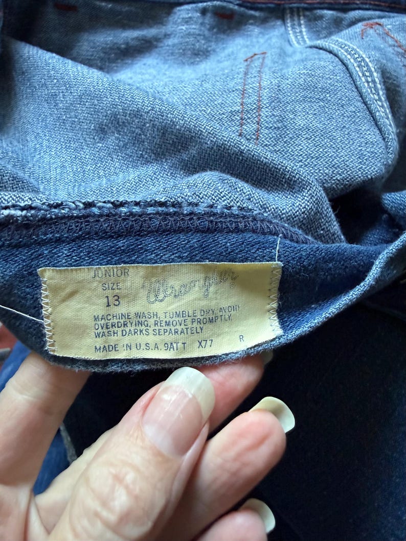 K&ouml;nnte beinhalten: Nahaufnahme einer blauen Jeans mit einem Wrangler-Etikett. Das Etikett lautet "Junior Gr&ouml;&szlig;e 13" und enth&auml;lt Waschanweisungen. Die Jeans wurde in den USA hergestellt.