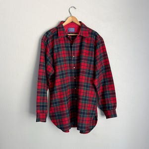 May include: A long-sleeve button-down shirt featuring a red, dark green, and navy blue plaid pattern. The shirt has a pointed collar, a front button placket, and buttoned cuffs. It is displayed on a wooden hanger against a plain white wall.
