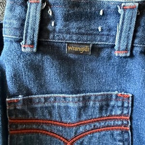 K&ouml;nnte beinhalten: Nahaufnahme einer blauen Jeans aus Denim mit orangefarbenen Nahtdetails. Das Wrangler-Etikett ist sichtbar. Die Ges&auml;&szlig;tasche hat ein markantes orangefarbenes Nahtmuster. Die Jeans hat G&uuml;rtelschlaufen und einen Knopfverschluss.