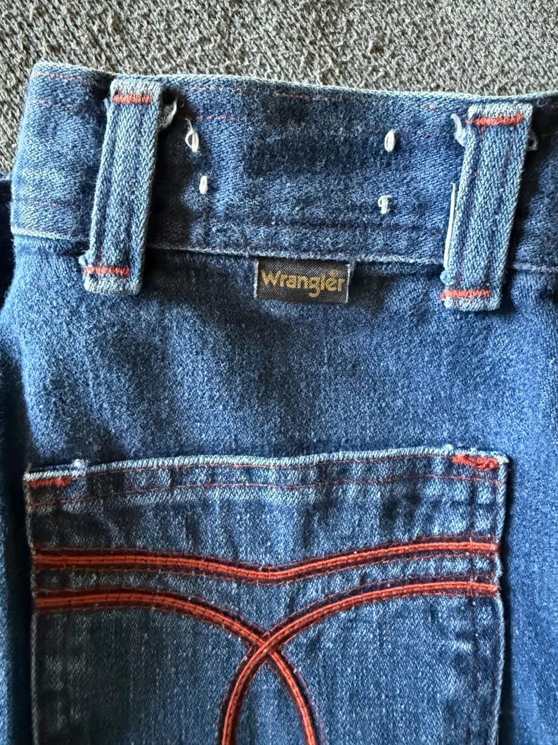 K&ouml;nnte beinhalten: Nahaufnahme einer blauen Jeans aus Denim mit orangefarbenen Nahtdetails. Das Wrangler-Etikett ist sichtbar. Die Ges&auml;&szlig;tasche hat ein markantes orangefarbenes Nahtmuster. Die Jeans hat G&uuml;rtelschlaufen und einen Knopfverschluss.