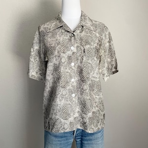 May include: A short-sleeved button-down shirt with a snakeskin pattern in shades of brown, tan, and white. The shirt has a classic collar, a chest pocket, and white buttons. The shirt is displayed on a mannequin with blue jeans.