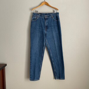 Peut inclure: Un jean bleu denim suspendu &agrave; un cintre en bois sur un mur blanc.
