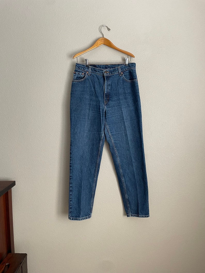 Peut inclure: Un jean bleu denim suspendu &agrave; un cintre en bois sur un mur blanc.