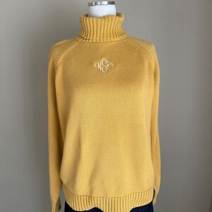 May include: Mustard yellow turtleneck jumper with long sleeves. The jumper features a ribbed turtleneck and hem, and a small embroidered design on the chest. The jumper is displayed on a mannequin.