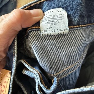 May include: A pair of dark blue denim jeans featuring a white interior care label with black text that reads 2183C 13, MACHINE WASH WARM NORMAL, ONLY NON-CHLORINE BLEACH IF NEEDED, TUMBLE DRY HOT, HOT IRON IF NEEDED, WASH AND DRY WITH LIKE COLORS, WPL 423, 219, 230 0702 19691-8, 15551-4837.