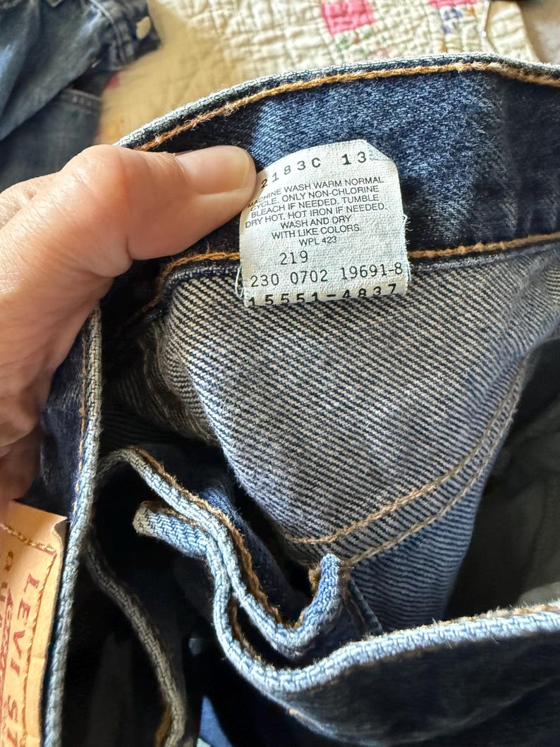 May include: A pair of dark blue denim jeans featuring a white interior care label with black text that reads 2183C 13, MACHINE WASH WARM NORMAL, ONLY NON-CHLORINE BLEACH IF NEEDED, TUMBLE DRY HOT, HOT IRON IF NEEDED, WASH AND DRY WITH LIKE COLORS, WPL 423, 219, 230 0702 19691-8, 15551-4837.