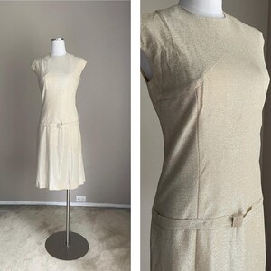 May include: A sleeveless, knee-length dress in a light beige color with a textured fabric. The dress has a fitted bodice and a slightly flared skirt. It features a simple design with a round neckline and a belt at the waist.