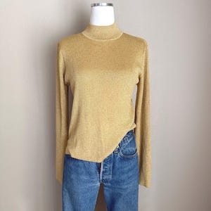 May include: A long-sleeved, ribbed mock neck sweater in a metallic gold finish, paired with classic blue denim jeans. The sweater features a fitted silhouette and a subtle shimmer, while the jeans have a traditional button-fly closure.