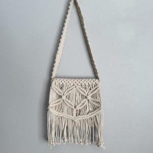 May include: Off-white macrame shoulder bag with a geometric design and fringe detail. The bag has a long strap and is made of woven cotton. The bag is hanging against a gray wall.
