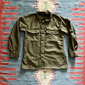 May include: Olive green long-sleeve shirt with two chest pockets and a button-down front. The shirt has a collared neckline and buttoned cuffs. The shirt is laid flat on a patterned rug with red, blue, and white stripes.