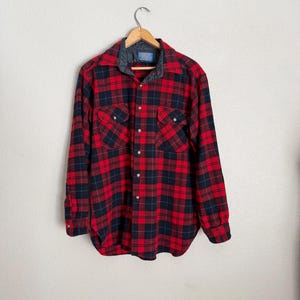 Puede incluir: Una camisa de franela a cuadros rojos y negros con cuello acolchado, cierre de botones y dos bolsillos en el pecho. La camisa est&aacute; colgada de una percha de madera contra una pared blanca.