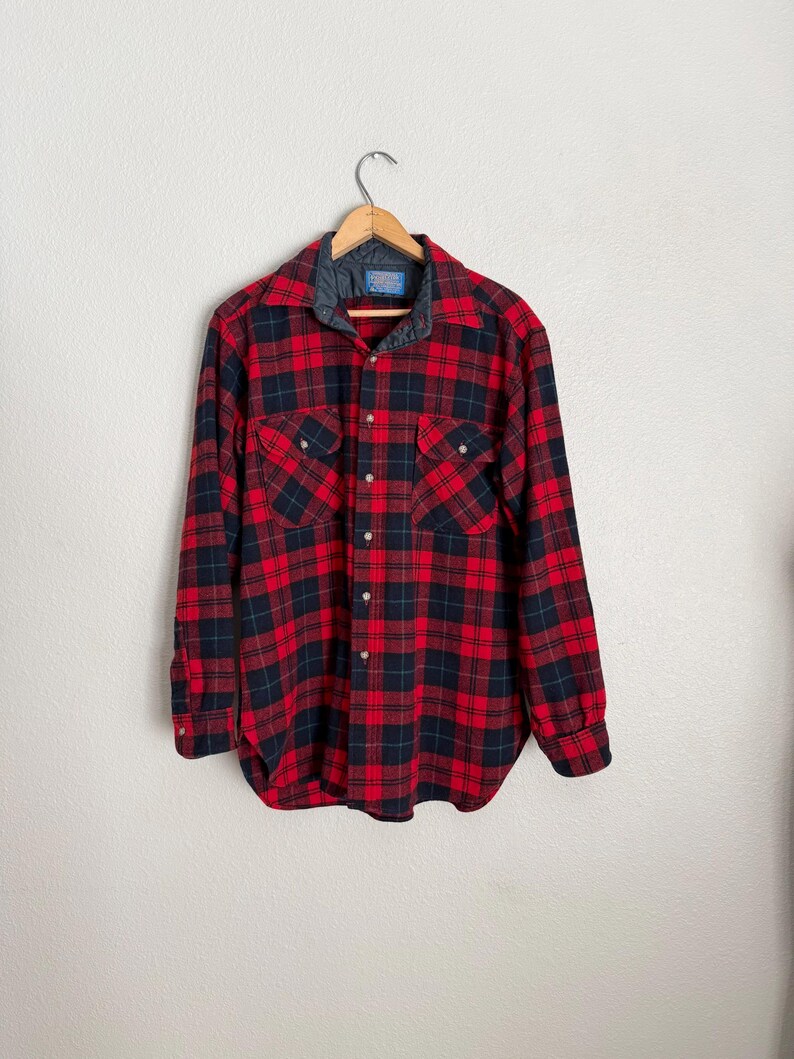 Puede incluir: Una camisa de franela a cuadros rojos y negros con cuello acolchado, cierre de botones y dos bolsillos en el pecho. La camisa est&aacute; colgada de una percha de madera contra una pared blanca.