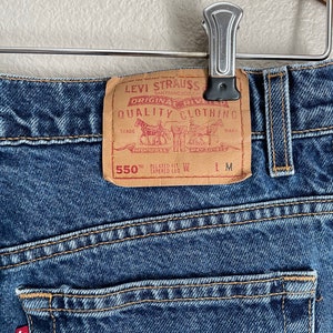 Peut inclure: Un jean en denim bleu avec un patch en cuir marron qui indique "Levi Strauss & Co. San Francisco, Cal. Original Rivelled Quality Clothing." Le patch comprend &eacute;galement un dessin d'une charrette tir&eacute;e par des chevaux et le texte "550th Relaxed Fit Tapered Leg".
