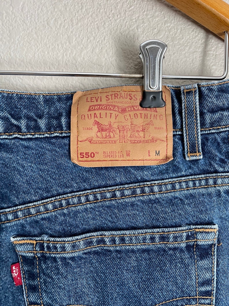 Peut inclure: Un jean en denim bleu avec un patch en cuir marron qui indique "Levi Strauss & Co. San Francisco, Cal. Original Rivelled Quality Clothing." Le patch comprend &eacute;galement un dessin d'une charrette tir&eacute;e par des chevaux et le texte "550th Relaxed Fit Tapered Leg".