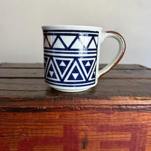 May include: A ceramic mug with a speckled off-white base and a dark blue geometric pattern featuring rows of triangles. The mug has a curved handle with a brown glazed finish and a cylindrical body with a slightly flared rim and a flat base.
