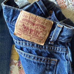 May include: A pair of blue denim jeans featuring a tan leather patch on the back waistband with the text Levi Strauss & Co. Original Riveted Quality Clothing Every Garment Guaranteed 550. The jeans include belt loops and a back pocket.