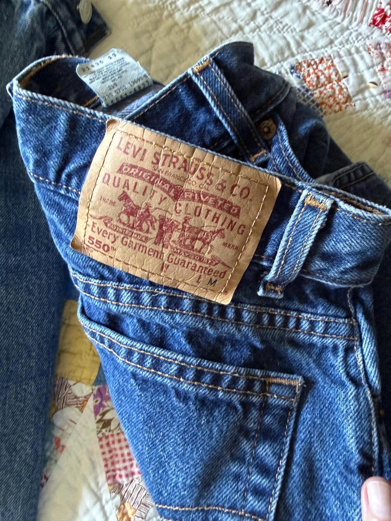 May include: A pair of blue denim jeans featuring a tan leather patch on the back waistband with the text Levi Strauss & Co. Original Riveted Quality Clothing Every Garment Guaranteed 550. The jeans include belt loops and a back pocket.