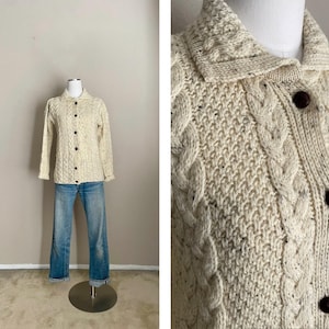 May include: A cream-colored cable knit cardigan with a button-up front and a collar. The cardigan has a speckled pattern and is made of a soft, cozy material.