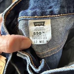 May include: Blue denim jeans with a white interior tag featuring the Levi's logo, 100% cotton, made in Mexico, 550 relaxed fit, tapered leg, and size 12 MIS M. The fabric has a visible twill weave and orange stitching along the waistband seams.