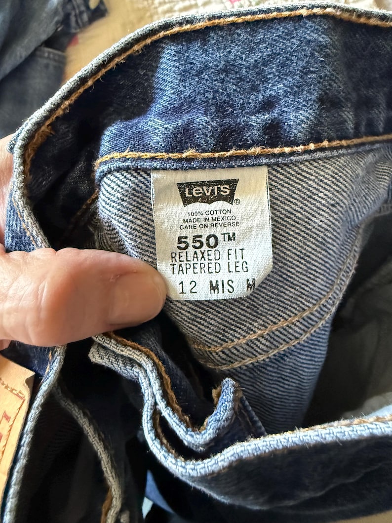 May include: Blue denim jeans with a white interior tag featuring the Levi's logo, 100% cotton, made in Mexico, 550 relaxed fit, tapered leg, and size 12 MIS M. The fabric has a visible twill weave and orange stitching along the waistband seams.