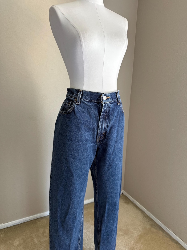 May include: High-waisted blue denim jeans featuring a classic five-pocket design, a metal button closure, and a zip fly. The jeans have a straight-leg cut with visible stitching along the seams and a medium-wash finish on the durable cotton fabric.
