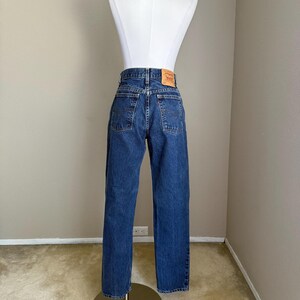 May include: A pair of dark blue denim jeans featuring a high-waisted fit, two back pockets with stitched arcuate detailing, a small red brand tab on the right pocket, and a tan leather-like patch on the back waistband displaying the Levi's logo.