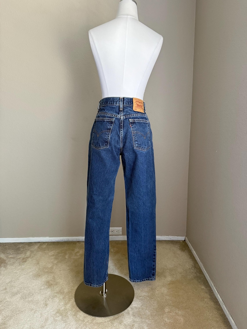 May include: A pair of dark blue denim jeans featuring a high-waisted fit, two back pockets with stitched arcuate detailing, a small red brand tab on the right pocket, and a tan leather-like patch on the back waistband displaying the Levi's logo.