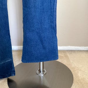 K&ouml;nnte beinhalten: Nahaufnahme einer blauen Jeans mit leicht ausgestelltem Bein. Die Jeans wird auf einem silbernen Metallst&auml;nder mit kreisf&ouml;rmiger Basis pr&auml;sentiert. Der Stoff hat eine strukturierte Optik.
