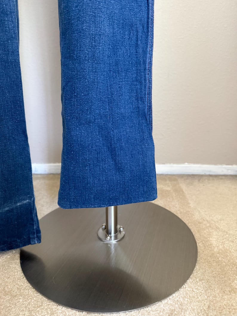 K&ouml;nnte beinhalten: Nahaufnahme einer blauen Jeans mit leicht ausgestelltem Bein. Die Jeans wird auf einem silbernen Metallst&auml;nder mit kreisf&ouml;rmiger Basis pr&auml;sentiert. Der Stoff hat eine strukturierte Optik.