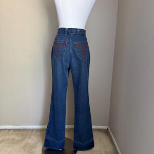 K&ouml;nnte beinhalten: Dunkelblaue Schlaghose aus Jeansstoff mit roten N&auml;hten auf den Ges&auml;&szlig;taschen. Die Jeans wird auf einer wei&szlig;en Schaufensterpuppe pr&auml;sentiert. Die Jeans hat eine hohe Taille und einen ausgestellten Bein-Design, typisch f&uuml;r die Mode der 1970er Jahre.